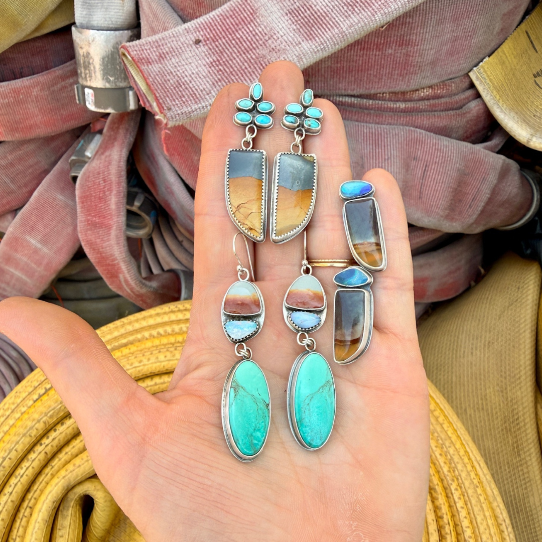 A hand holding a set of turquoise and stone jewelry, including dangle earrings and matching rings, against a background of pinkish-brown fabric and yellow woven material.